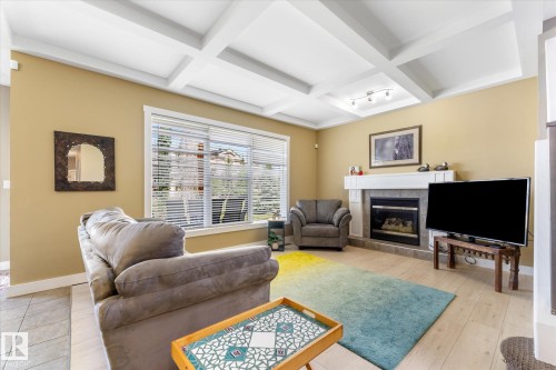 Living room featuring a coffered ceiling, a large window with white blinds, wood-finish flooring, a fireplace with a stone surround, and a white mantel - 488 Macewan Road, Edmonton, AB 