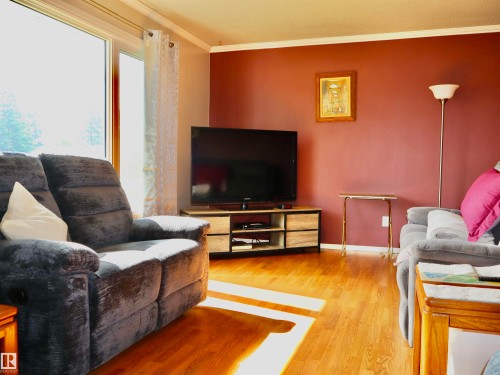 Living area featuring wood-finish flooring, a large window, a media console with open shelving, and a wall with a deep red accent - 5530 46 Street, Barrhead, AB 