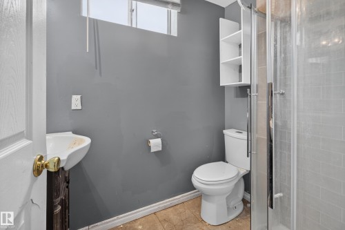 Compact bathroom featuring a white toilet, glass-enclosed shower, white wall-mounted shelving unit, and a white basin sink with a dark wood-finish vanity - 9315 132A Avenue, Edmonton, AB 