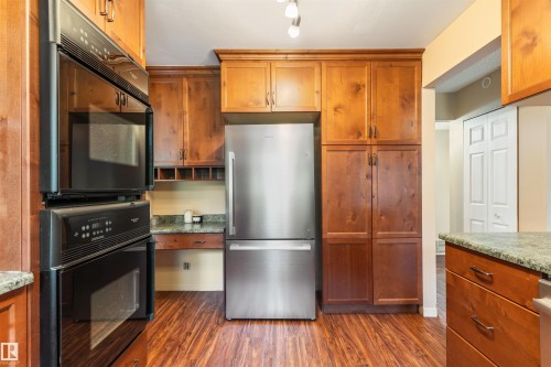 Kitchen featuring wood-finish flooring, full-height wood cabinetry, stainless steel refrigerator, built-in double ovens, and stone countertops - 20515 5 Avenue Sw, Edmonton, AB 