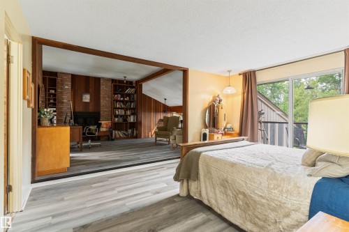 Spacious room featuring light wood-finish flooring, a sliding glass door to an exterior balcony, and an open doorway to an adjacent room with wood paneling and a brick fireplace - 20515 5 Avenue Sw, Edmonton, AB 