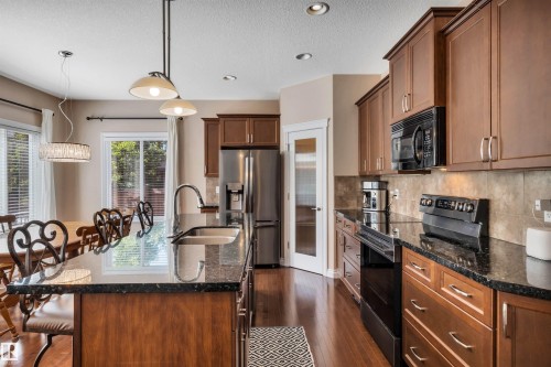 Contemporary kitchen featuring an expansive island with a double basin sink, dark stone countertops, wood-finish cabinetry, stainless steel appliances, and recessed lighting - 18 Voleta Crest, Spruce Grove, AB 