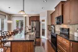 Contemporary kitchen featuring an expansive island with a double basin sink, dark stone countertops, wood-finish cabinetry, stainless steel appliances, and recessed lighting - 