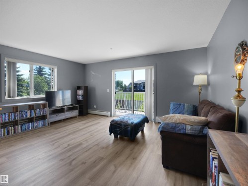 Spacious living area featuring light wood-finish flooring, a large window overlooking trees, and a glass sliding door opening to a private balcony - #217 240 Spruce Ridge Rd, Spruce Grove, AB - Indoor Photo Showing Living Room