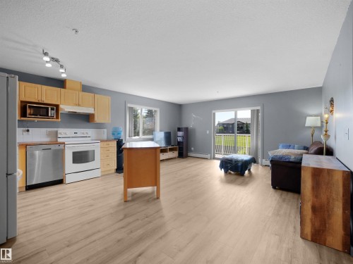 Open-concept living area featuring wood-finish flooring, a kitchen with light wood cabinetry, white appliances, and a stainless steel dishwasher - #217 240 Spruce Ridge Rd, Spruce Grove, AB - Indoor Photo Showing Kitchen