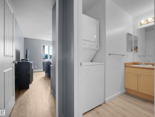 Compact laundry area featuring a stacked washer and dryer, adjacent to a bathroom with a single vanity, wood-finish flooring, and recessed medicine cabinet - #217 240 Spruce Ridge Rd, Spruce Grove, AB - Indoor Photo Showing Laundry Room