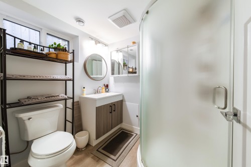 Contemporary bathroom featuring a curved glass shower enclosure, a vanity with wood-finish cabinetry, and a circular wall mirror - 10811 73 Avenue, Edmonton, AB 