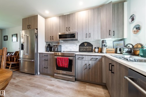 Modern kitchen featuring wood-finish cabinetry with black hardware, stainless steel appliances, light-toned countertops, a tiled backsplash, and wood-finish flooring - 10811 73 Avenue, Edmonton, AB 