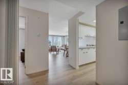 Light wood-finish flooring extending into the living area, white kitchen cabinetry with dark hardware, a dining area, and a bright living space with large windows - 