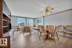 Open-concept living area featuring light wood-finish flooring, multiple windows with vertical blinds, a ceiling fan with integrated lighting, and a built-in wood shelving unit - 