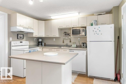 Kitchen featuring a central island with a solid surface countertop, white cabinetry with silver-tone hardware, wood-finish flooring, and white appliances including a range, refrigerator, and dishwasher - 171 Ridgewood, St. Albert, AB 