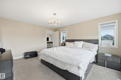 Spacious bedroom featuring light neutral wall tones, plush carpet flooring, a contemporary chandelier, and multiple windows with roller blinds - 76 Jacobs Close, St. Albert, AB 