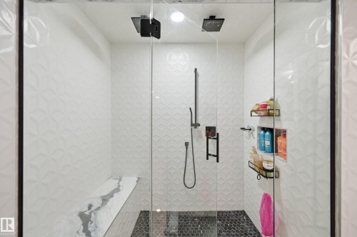 Glass-enclosed shower featuring dual matte black rain shower heads, a wall-mounted hand shower, a built-in bench with a stone-finish surface, patterned white tile walls, and a black hex tile floor - 76 Jacobs Close, St. Albert, AB 