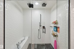 Glass-enclosed shower featuring dual matte black rain shower heads, a wall-mounted hand shower, a built-in bench with a stone-finish surface, patterned white tile walls, and a black hex tile floor - 
