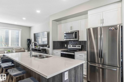 Kitchen featuring a central island with an undermount double sink and chrome faucet, white cabinetry, stainless steel appliances, a dark herringbone tile backsplash, and recessed lighting - 1908 25 Street, Edmonton, AB 