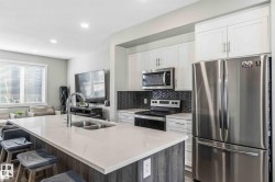 Kitchen featuring a central island with an undermount double sink and chrome faucet, white cabinetry, stainless steel appliances, a dark herringbone tile backsplash, and recessed lighting - 