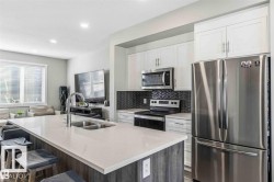 Open-concept kitchen featuring a large island with an undermount double-basin sink, stainless steel appliances, white cabinetry, and a chevron pattern tile backsplash - 
