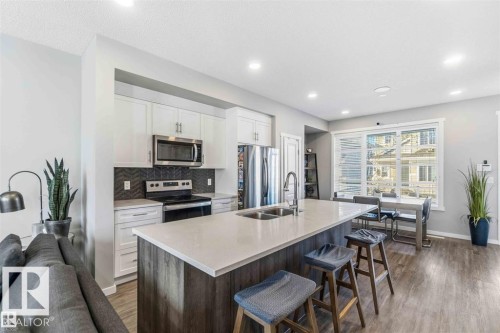 Modern kitchen with stainless steel appliances, white cabinetry, a dark herringbone tile backsplash, and a large island with an integrated double sink - 1908 25 Street, Edmonton, AB 
