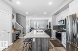 Contemporary kitchen featuring an oversized island with a built-in sink, stainless steel appliances, white cabinetry, and a dark herringbone tile backsplash - 