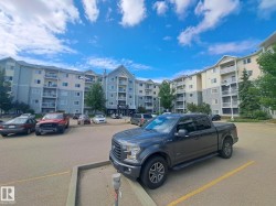 Multi-story residential building featuring balconies with metal railings, a central entrance with a canopy, and a parking lot with marked spaces - 