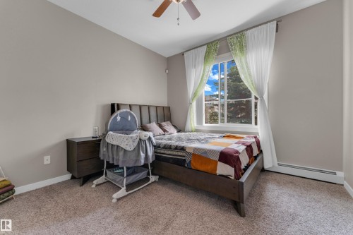 Carpeted bedroom featuring a large window, a wood-finish bed frame with a paneled headboard, and a ceiling fan - 115 812 Welsh Drive Sw, Edmonton, AB 