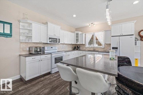 Modern kitchen featuring a central island with a stone-finish countertop, white cabinetry, integrated appliances, wood-finish flooring, and a tile backsplash - 4708 47A Avenue, Onoway, AB 