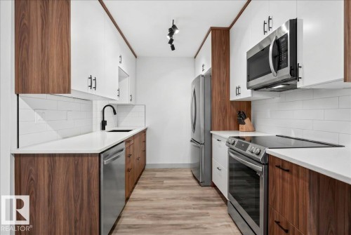 Galley kitchen featuring wood-finish lower cabinetry, white upper cabinetry, stainless steel appliances, white subway tile backsplash, and light wood-finish flooring - Edmonton, AB 