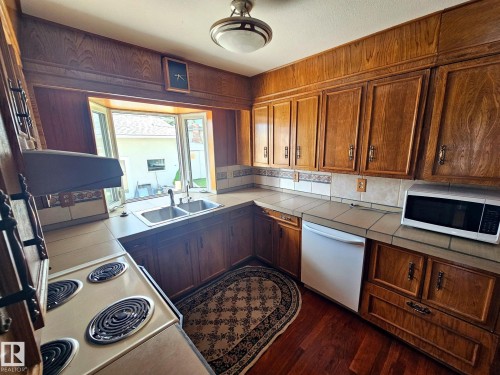 Kitchen featuring extensive wood cabinetry, a double basin stainless steel sink, a tile countertop, a tile backsplash, and wood-finish flooring - 7612 40 Avenue Nw, Edmonton, AB - Indoor Photo Showing Kitchen With Double Sink
