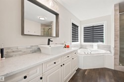 Spacious bathroom featuring a built-in corner soaking tub, dual windows with blinds, a vanity with a rectangular vessel sink, quartz-finish countertop, and wood-finish flooring - 