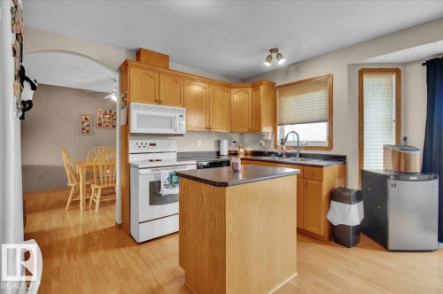 Kitchen featuring wood-finish flooring, light wood cabinetry, a central island with a dark countertop, white appliances, and a window above the sink - 5506 53St, Mundare, AB 
