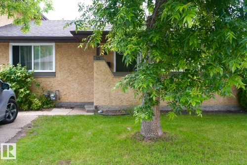 Tan stucco exterior featuring a large front window, dark trim, and a mature shade tree - 25 8930 99 Avenue, Fort Saskatchewan, AB 