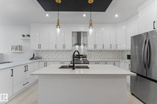 Contemporary kitchen featuring an island with an undermount sink and matte black faucet, extensive white cabinetry with black hardware, stone countertops, and a patterned tile backsplash - 3606 42 Avenue, Beaumont, AB 