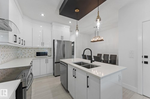 Modern kitchen featuring white cabinetry, stainless steel appliances, a white quartz countertop island with a double basin sink, and a decorative tile backsplash - 3606 42 Avenue, Beaumont, AB 