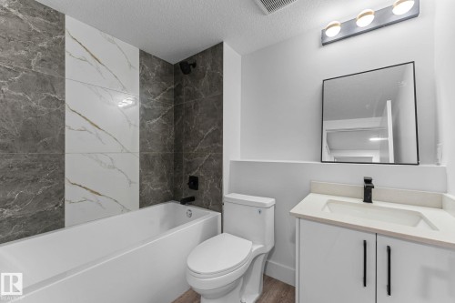 Bathroom featuring a soaking tub with contrasting tile surround, white vanity with integrated sink, modern black fixtures, and wood-finish flooring - 3606 42 Avenue, Beaumont, AB 