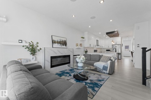 Open-concept living area featuring a modern fireplace with a marble-look surround, light wood-finish flooring, recessed lighting, white floating shelves, and a contemporary kitchen with white cabinetry and stainless steel appliances - 3606 42 Avenue, Beaumont, AB 
