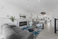 Open-concept living area featuring a modern fireplace with a marble-look surround, light wood-finish flooring, recessed lighting, white floating shelves, and a contemporary kitchen with white cabinetry and stainless steel appliances - 