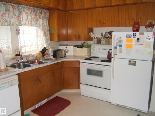 Kitchen featuring wood cabinetry, a dual basin stainless steel sink, and white appliances - 4606 51 Avenue, Two Hills, AB 