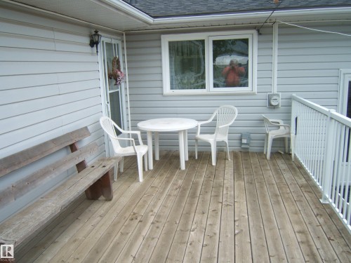 Wood-finish deck featuring light grey horizontal siding, white railing, and an outdoor wall sconce - 4606 51 Avenue, Two Hills, AB 