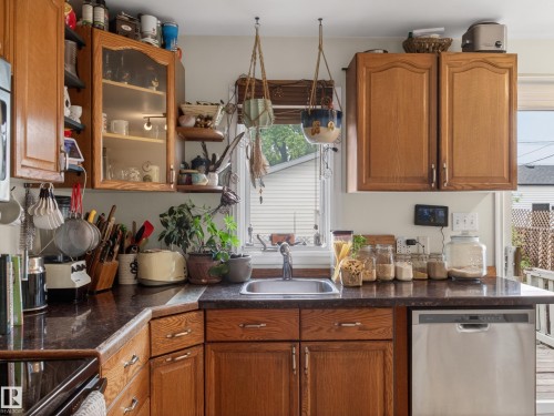 Kitchen featuring wood cabinetry, dark countertops, a stainless steel sink, a stainless steel dishwasher, and a window with blinds - 10809 150 Street, Edmonton, AB 