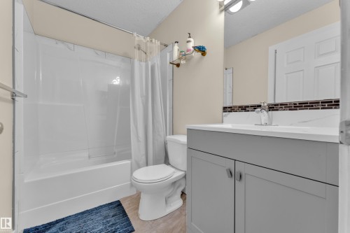Bathroom featuring a white bathtub/shower combination with a textured surround, a toilet, and a vanity with a light gray cabinet base and a white countertop - 2362 29A Avenue, Edmonton, AB 