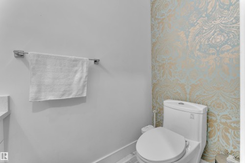 Restroom featuring a wall with gold and light blue patterned wallpaper, a white toilet, and a chrome towel bar - 2630 16A Avenue, Edmonton, AB 
