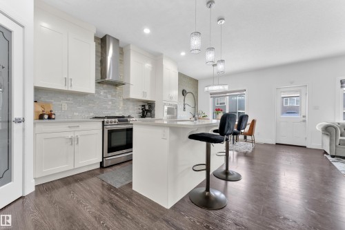 Modern kitchen featuring white shaker cabinetry, stainless steel appliances, a light-toned backsplash, a central island with countertop seating, and wood-finish flooring - 2630 16A Avenue, Edmonton, AB 