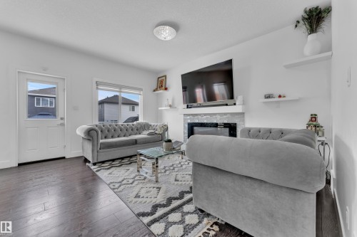 Living area featuring dark wood-finish flooring, a contemporary ceiling light fixture, a fireplace with a light-toned tile surround and white mantel, and a window with white trim - 2630 16A Avenue, Edmonton, AB 