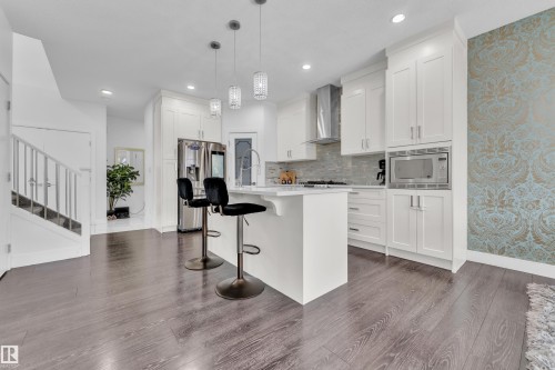 Modern kitchen featuring a central island with seating, white shaker cabinetry, stainless steel appliances, and wood-finish flooring - 2630 16A Avenue, Edmonton, AB 