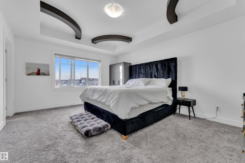 Spacious room featuring tray ceilings with dark architectural accents, a large window providing ample natural light, light gray carpeting, and white walls - 2630 16A Avenue, Edmonton, AB 