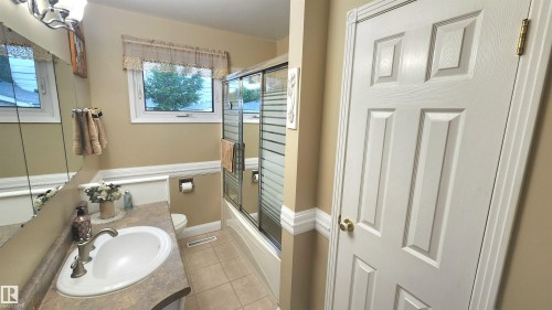 Bathroom featuring a vanity with an oval basin, a window above the toilet, a tub with a sliding glass shower door, and tile flooring - 13307 Delwood Road, Edmonton, AB 