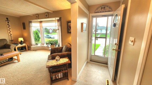 Entryway with a solid white door and decorative glass insert, opening to a living area featuring a large bay window, textured carpeting, and exposed ceiling beams - 13307 Delwood Road, Edmonton, AB 
