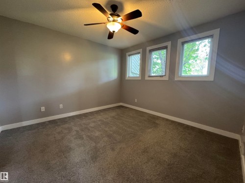 Spacious room featuring three rectangular windows with white trim, a ceiling fan with integrated lighting, textured neutral carpeting, and light gray wall paint - 6441 180 Street, Edmonton, AB 
