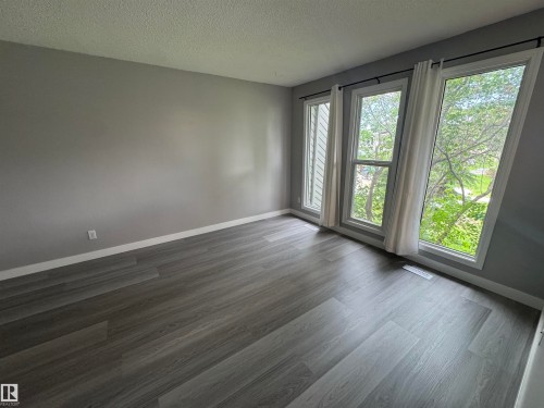 Room featuring wood-finish flooring, neutral gray walls, white baseboards, and a series of three large windows providing ample illumination - 6441 180 Street, Edmonton, AB 