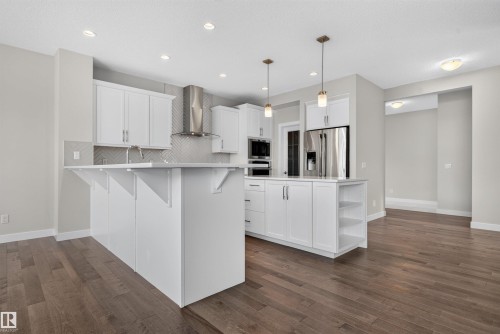Contemporary kitchen featuring white cabinetry, stainless steel appliances, a herringbone tile backsplash, an island with built-in shelving, and wood-finish flooring - 9432 229 Street, Edmonton, AB 